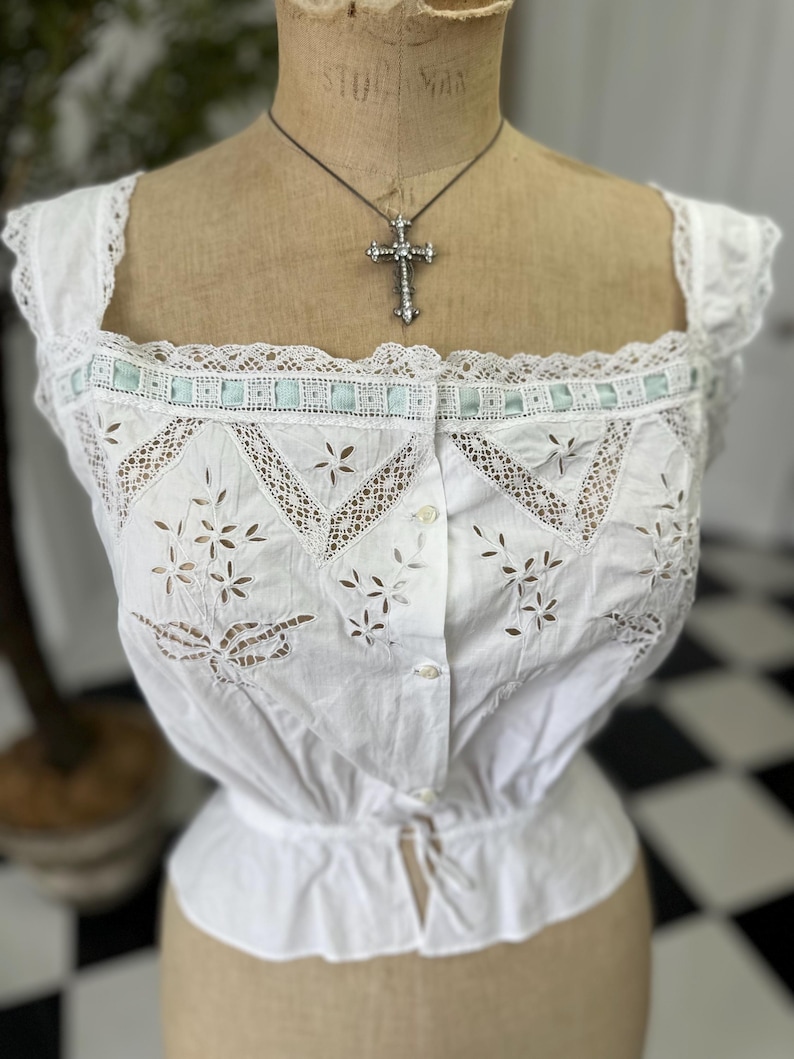 Puede incluir: Una blusa de algod&oacute;n blanco con delicado ribete de encaje y detalles florales bordados. La blusa tiene un frente abotonado y una cintura ajustada.