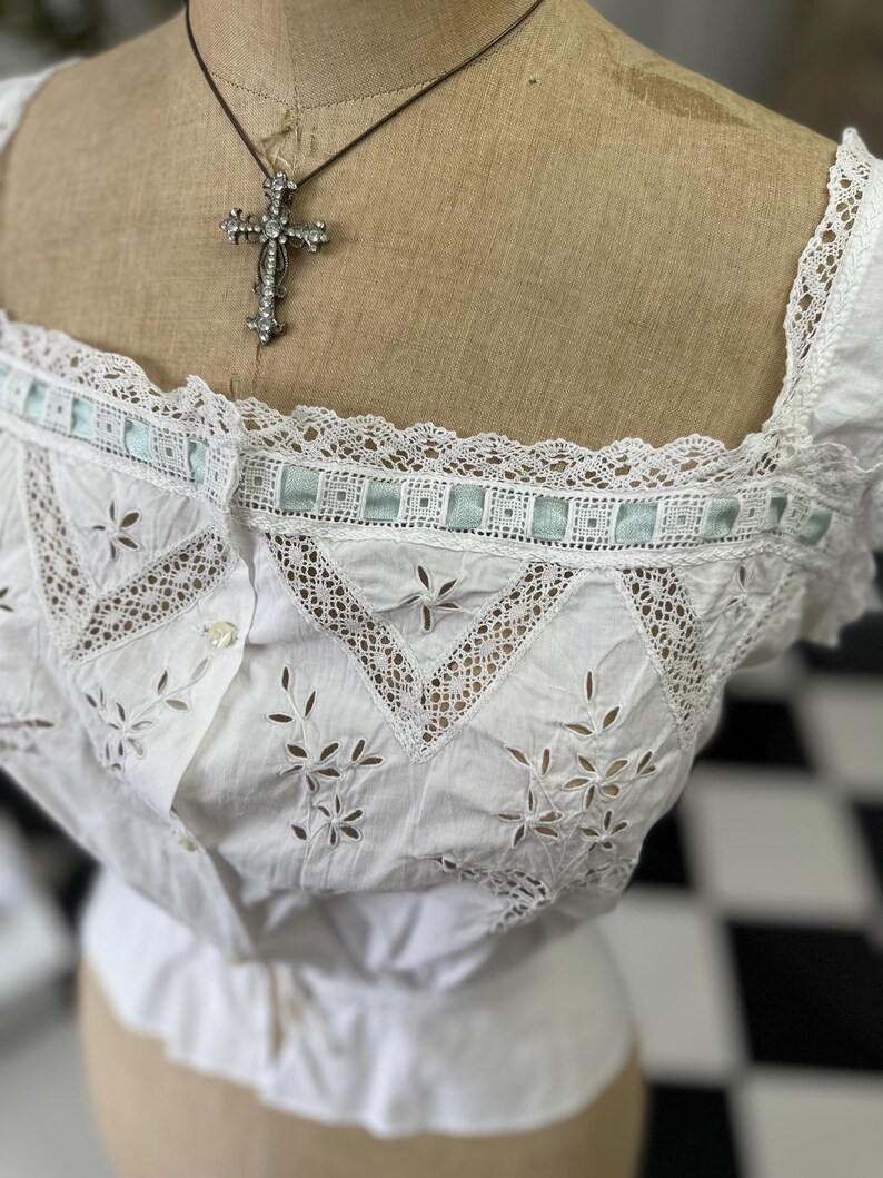 Puede incluir: Blusa blanca de algod&oacute;n con delicado ribete de encaje y detalles bordados. La blusa tiene un escote cuadrado y un cierre de botones en la parte delantera. Un collar de cruz plateada es visible en el maniqu&iacute;.