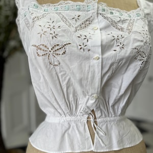 Puede incluir: Blusa blanca de algod&oacute;n con delicado encaje y bordados intrincados. La blusa tiene una cintura fruncida y un frente abotonado.
