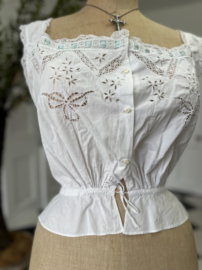 Puede incluir: Blusa blanca de algod&oacute;n con delicado encaje y bordados intrincados. La blusa tiene una cintura fruncida y un frente abotonado.