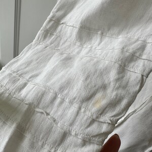 May include: White linen fabric with a subtle texture and a small yellow stain. The fabric has multiple rows of stitching.