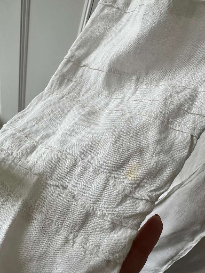 May include: White linen fabric with a subtle texture and a small yellow stain. The fabric has multiple rows of stitching.