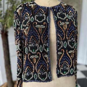 Könnte beinhalten: Eine schwarze Perlenjacke mit Paisley-Muster in Blau-, Grün- und Goldtönen. Die Jacke hat einen vorderen Öffnung und lange Ärmel.