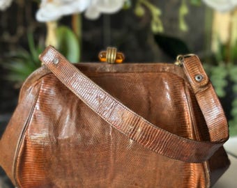 Könnte beinhalten: Eine braune Vintage-Handtasche mit einer strukturierten, reptilienartigen Oberfläche. Die Tasche hat einen passenden Riemen und einen dekorativen Verschluss mit bernsteinfarbenen Akzenten. Die Tasche liegt auf einer weißen Oberfläche.