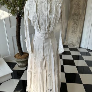 May include: A white vintage dress with delicate embroidery and lace detailing. The dress has a high neck and long sleeves, and the skirt is full and flowing.