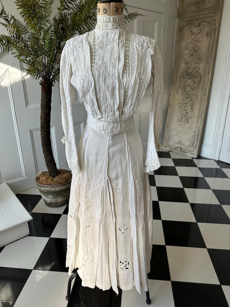 May include: A white vintage dress with delicate embroidery and lace detailing. The dress has a high neck and long sleeves, and the skirt is full and flowing.