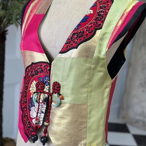 Puede incluir: Un chaleco sin mangas hecho de tela de patchwork en tonos de rosa, dorado y verde. El chaleco presenta un dise&ntilde;o floral decorativo y un cierre de corbata con borla.