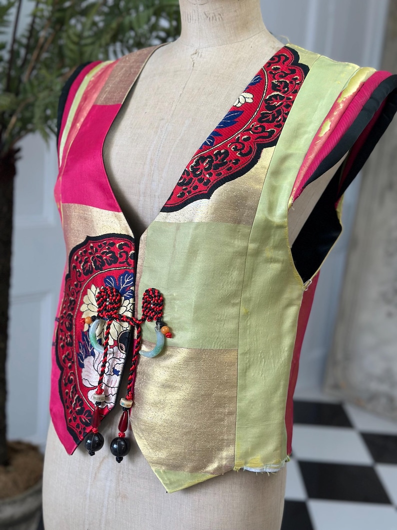 Puede incluir: Un chaleco sin mangas hecho de tela de patchwork en tonos de rosa, dorado y verde. El chaleco presenta un dise&ntilde;o floral decorativo y un cierre de corbata con borla.