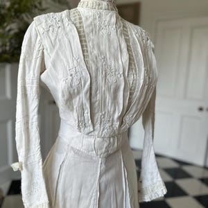May include: A white vintage dress with long sleeves and intricate lace detailing. The dress has a high neck and a fitted bodice with a gathered skirt.