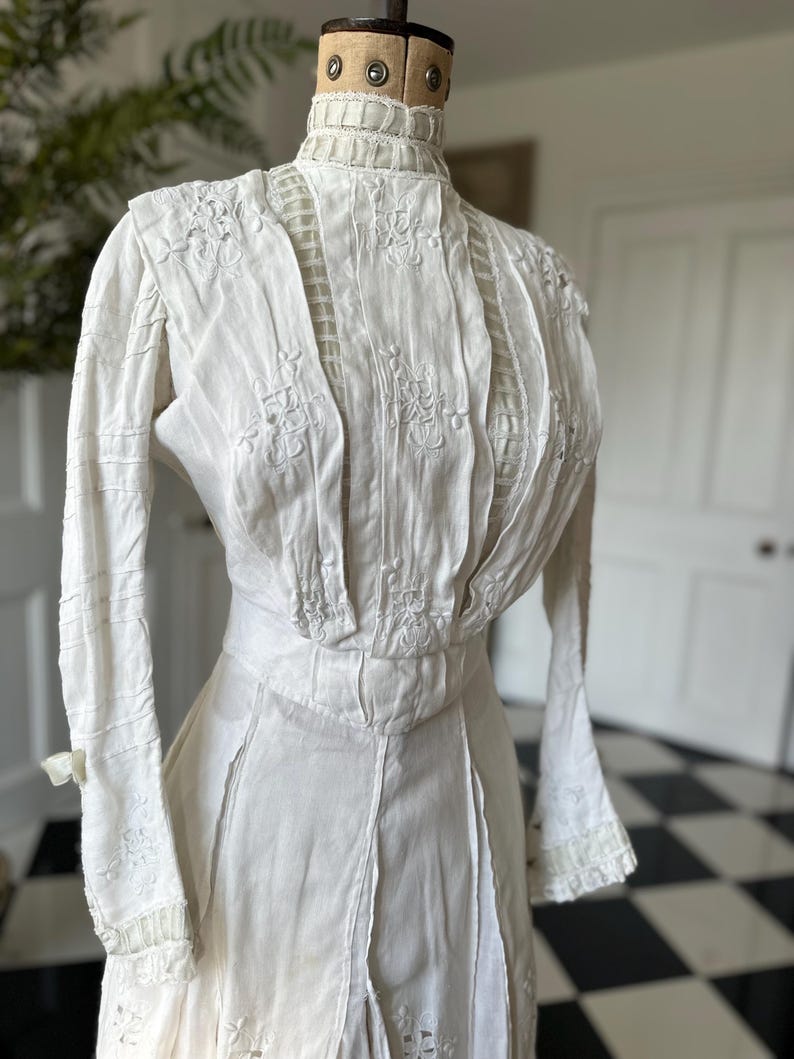 May include: A white vintage dress with long sleeves and intricate lace detailing. The dress has a high neck and a fitted bodice with a gathered skirt.