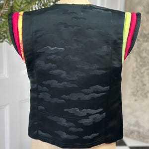 Puede incluir: Chaleco negro sin mangas con un patr&oacute;n de nubes y ribete de colores. El ribete es una combinaci&oacute;n de rosa, amarillo y verde.