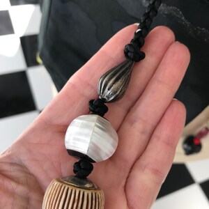 Puede incluir: Un collar de cord&oacute;n negro con tres cuentas. Las cuentas son una l&aacute;grima de plata, una cuenta redonda blanca y negra y una cuenta redonda marr&oacute;n y blanca.