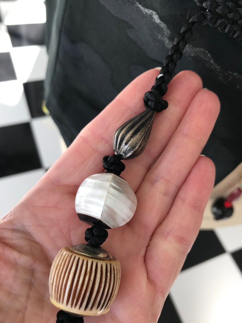 Puede incluir: Un collar de cord&oacute;n negro con tres cuentas. Las cuentas son una l&aacute;grima de plata, una cuenta redonda blanca y negra y una cuenta redonda marr&oacute;n y blanca.