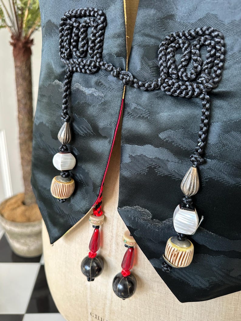 Puede incluir: Un chaleco de seda negro con intrincados detalles de cord&oacute;n negro y cuentas decorativas. El chaleco tiene un ribete rojo y dorado y presenta un dise&ntilde;o &uacute;nico con un patr&oacute;n de nubes.