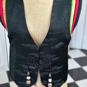Puede incluir: Un chaleco de seda negro con estampado de nubes y cierre de borla decorativo. El chaleco tiene ribetes rojos, amarillos y verdes en los hombros.