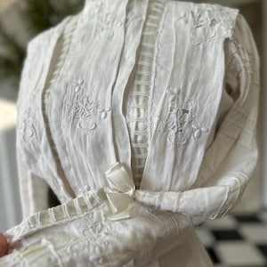 May include: A white vintage blouse with a high neck and long sleeves. The blouse is made of a delicate fabric and features intricate embroidery and lace details.