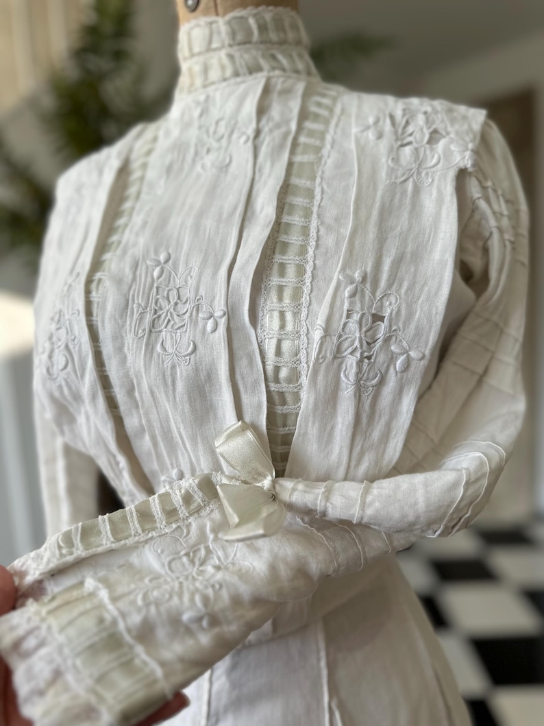 May include: A white vintage blouse with a high neck and long sleeves. The blouse is made of a delicate fabric and features intricate embroidery and lace details.