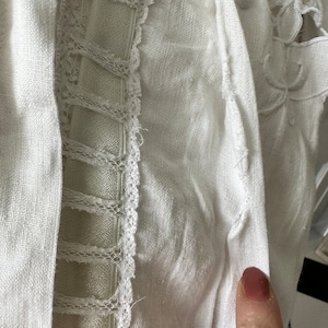 May include: White linen fabric with a delicate lace trim. The fabric has a slightly textured surface and is folded over on itself.