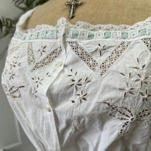 Puede incluir: Top blanco de algod&oacute;n con delicado ribete de encaje y detalles florales bordados. El top tiene un frente abotonado y una cintura fruncida.