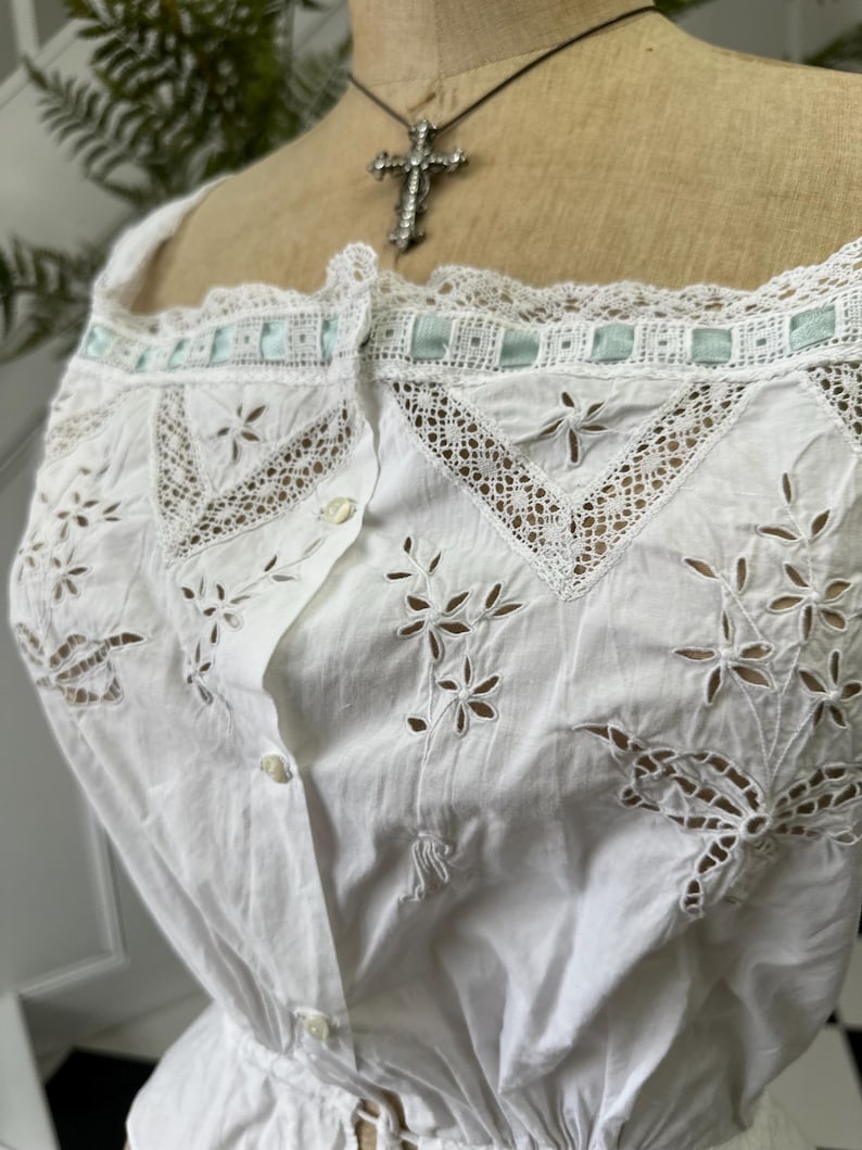 Puede incluir: Top blanco de algod&oacute;n con delicado ribete de encaje y detalles florales bordados. El top tiene un frente abotonado y una cintura fruncida.