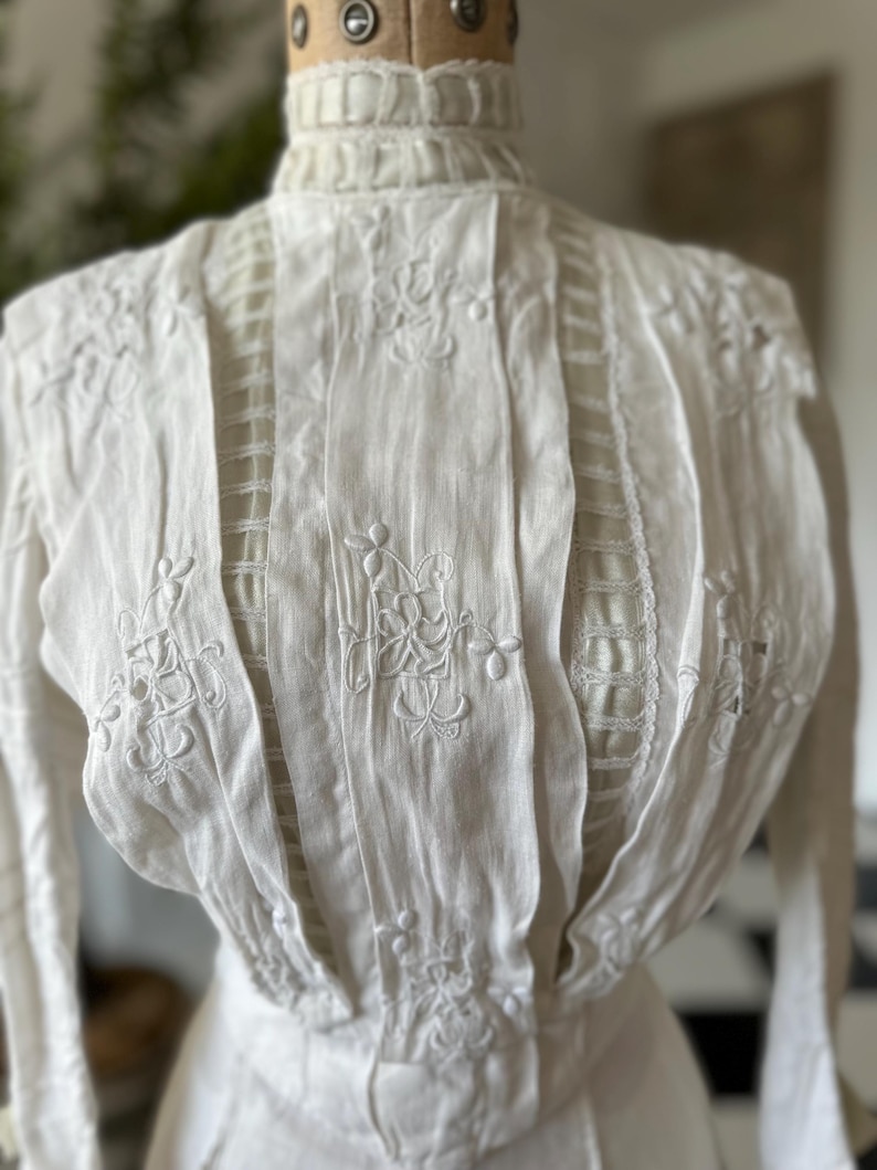 May include: White cotton blouse with a high neck and intricate embroidered floral details. The blouse has a fitted bodice and long sleeves.
