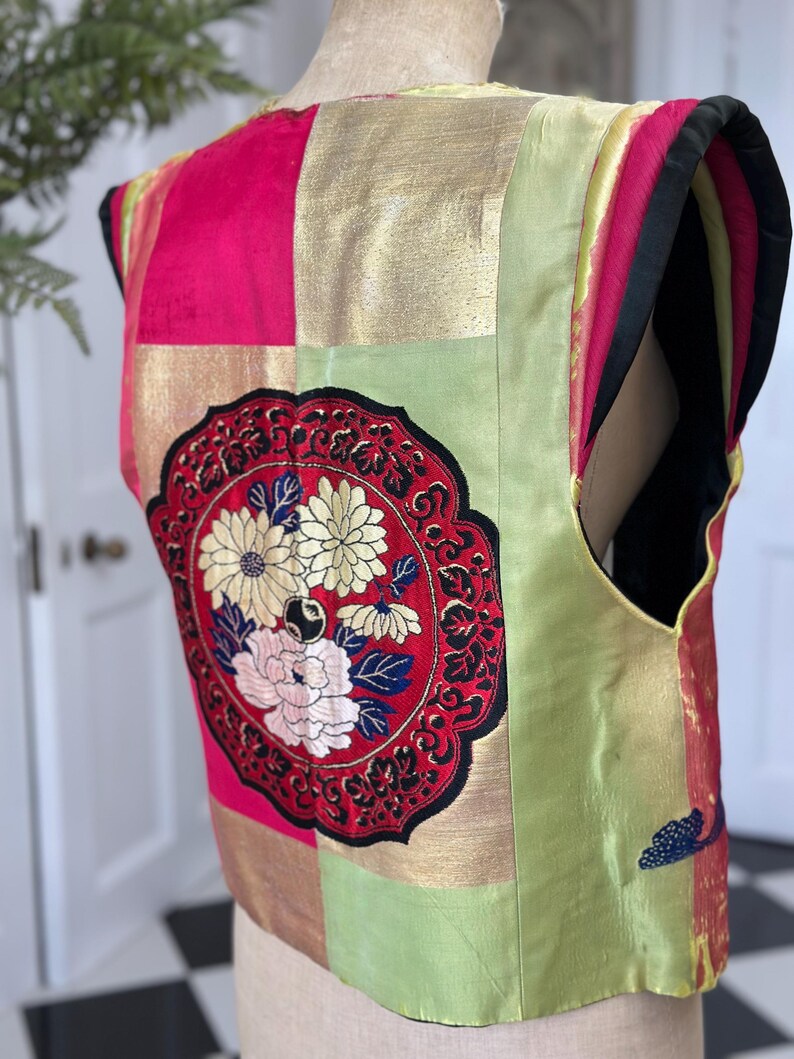 Puede incluir: Un chaleco sin mangas hecho de tela de retazos en tonos rojos, dorados y verdes. El chaleco presenta un gran c&iacute;rculo bordado con dise&ntilde;os florales en negro, blanco y azul.