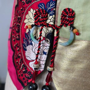 Puede incluir: Una chaqueta de estilo chino roja y dorada con un borla negro y rojo y un colgante de jade. La chaqueta tiene un patr&oacute;n floral y est&aacute; hecha de seda.