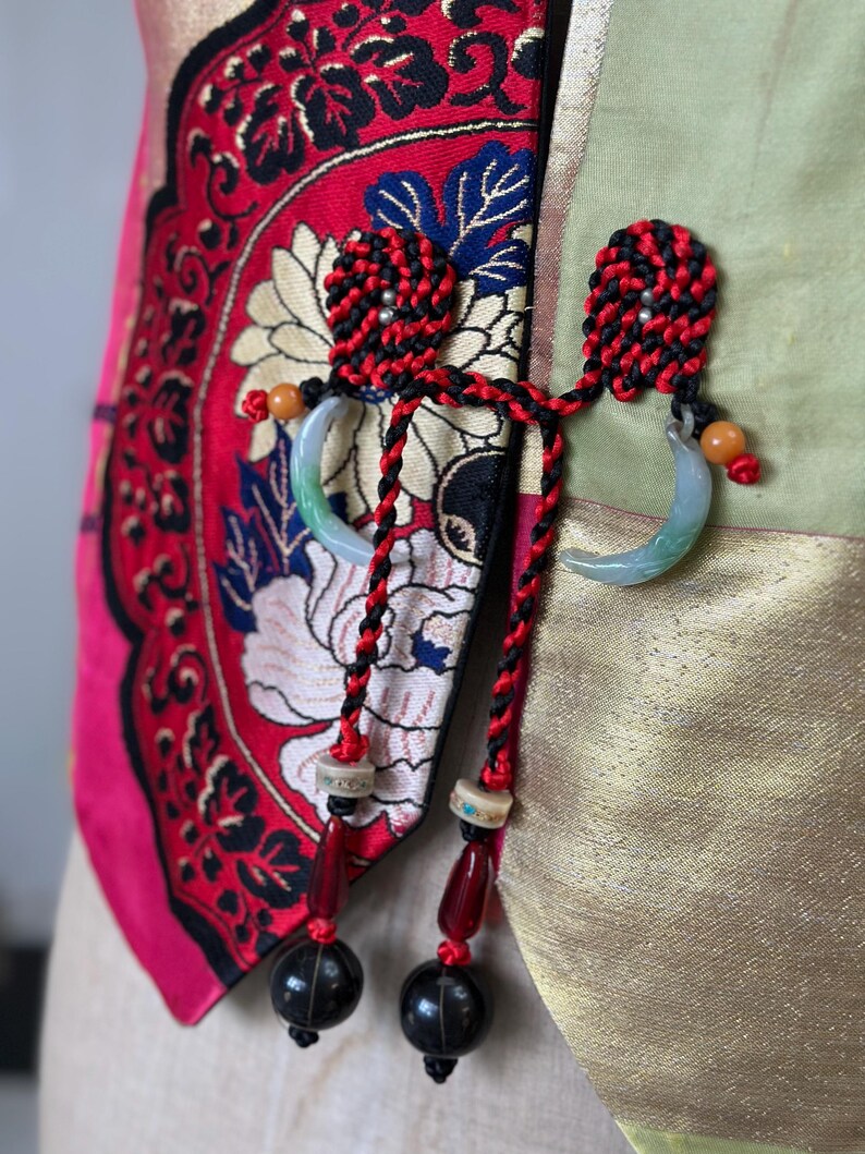 Puede incluir: Una chaqueta de estilo chino roja y dorada con un borla negro y rojo y un colgante de jade. La chaqueta tiene un patr&oacute;n floral y est&aacute; hecha de seda.