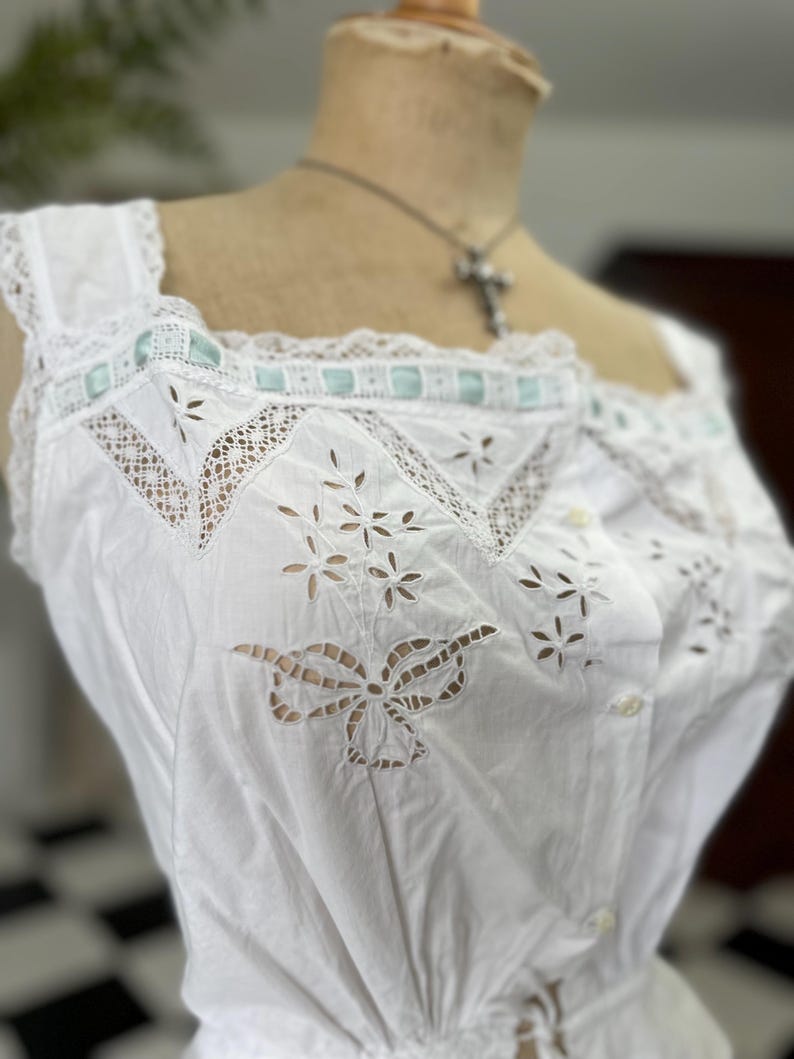 Puede incluir: Camis&oacute;n de algod&oacute;n blanco con encaje de ojal delicado y un ribete de cinta azul claro. El camis&oacute;n tiene cierre de bot&oacute;n y cintura fruncida.