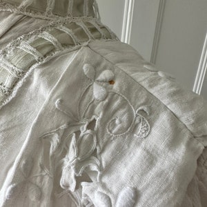 May include: White linen fabric with delicate embroidered floral designs. The fabric has a slightly textured surface and is accented with a delicate lace trim.