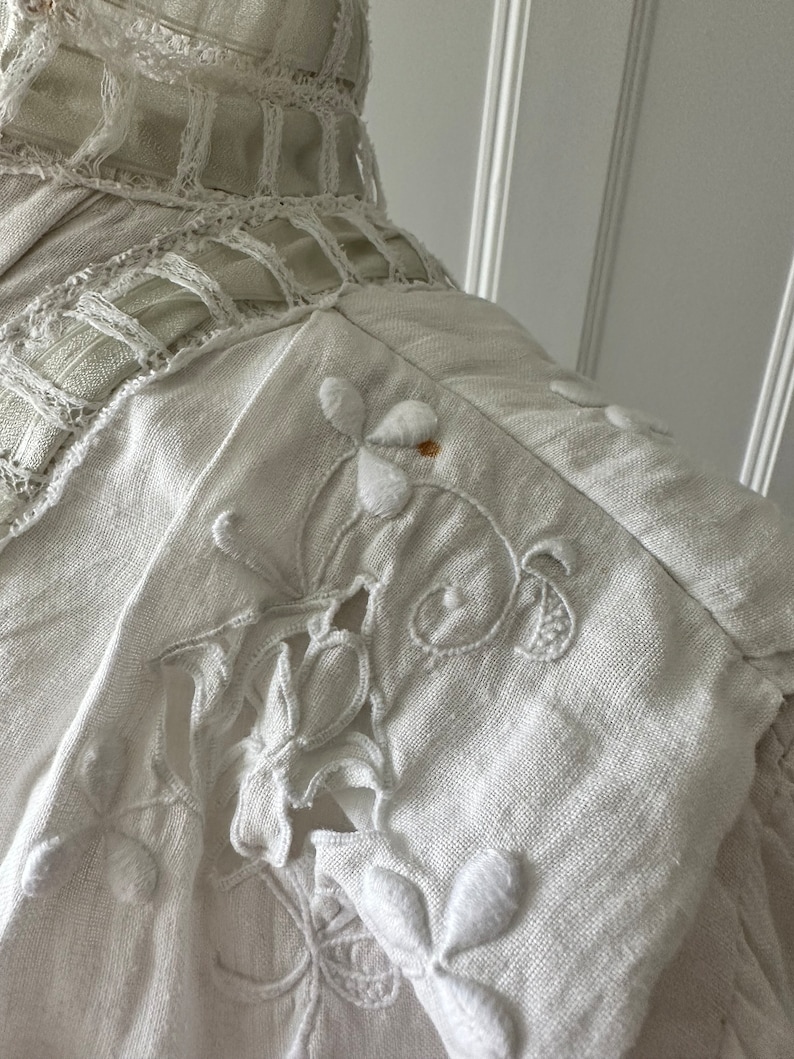 May include: White linen fabric with delicate embroidered floral designs. The fabric has a slightly textured surface and is accented with a delicate lace trim.