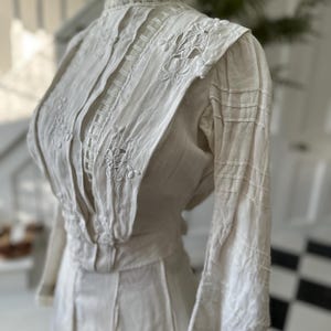 May include: A white vintage dress with a high collar and long sleeves. The dress is made of a lightweight fabric and features delicate lace and embroidery details.