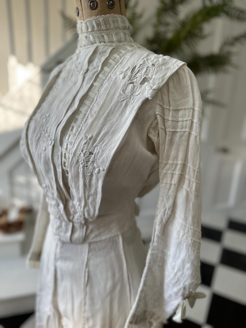 May include: A white vintage dress with a high collar and long sleeves. The dress is made of a lightweight fabric and features delicate lace and embroidery details.