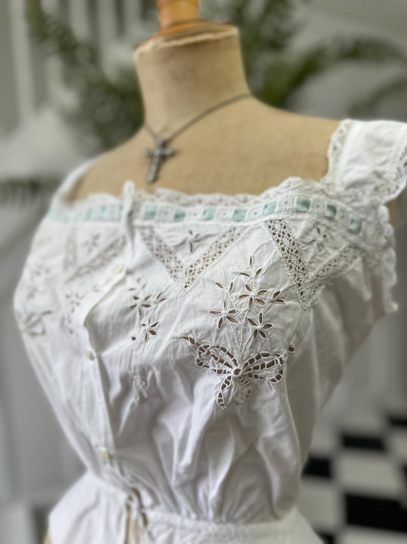 Puede incluir: Top blanco de algod&oacute;n con delicado encaje y bordados intrincados. El top tiene un escote cuadrado y un cierre de botones en la parte delantera.