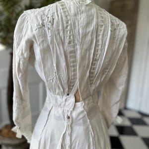 May include: A white cotton dress with a high neck and long sleeves. The dress is decorated with delicate lace and embroidery. The back of the dress features a unique open back design.