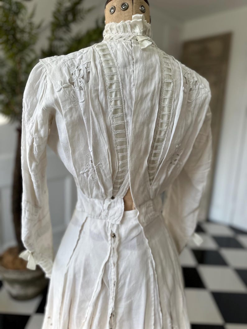 May include: A white cotton dress with a high neck and long sleeves. The dress is decorated with delicate lace and embroidery. The back of the dress features a unique open back design.