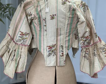 May include: A vintage, cream-colored, striped and floral patterned jacket with pink and green accents. The jacket has long sleeves with ruffled cuffs and a pointed hemline. The jacket is displayed on a dress form.