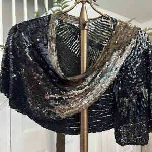 May include: A black sequined bolero jacket with a draped neckline. The jacket features a mix of black, silver, and bronze sequins, creating a shimmering effect. The garment is displayed on a gold-colored hanger.