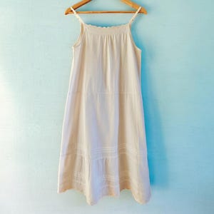 以下が含まれることがあります： 淡いクリーム色のスリップドレスが、薄い青色の壁を背景に木製のハンガーに掛けられています。ドレスは、細いスパゲッティストラップ、スカラップのネックライン、ティアードレーストリムの裾が特徴です。ドレスは軽量の生地で作られています。