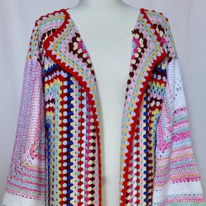 May include: A long, open-front crochet cardigan in a vibrant patchwork of colors. The design features a red trim and a mix of pinks, blues, yellows, and whites. The sleeves are long and flared, and the cardigan is displayed on a mannequin.