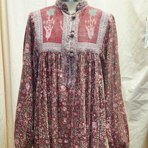 Puede incluir: Vestido largo burdeos con estampado floral y mangas largas. El vestido presenta un cuello alto con botones y paneles decorativos en el corpiño. El dobladillo y los puños tienen un borde contrastante. El vestido se exhibe en un maniquí.
