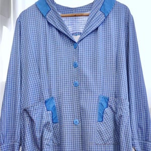 May include: A vintage blue and white checked dress with a collar and button-down front. The dress has two front pockets with blue accents and a small embroidered detail near the hem. The dress is hanging on a wooden hanger.
