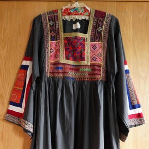 Puede incluir: Vestido gris oscuro con detalles bordados coloridos. El corpiño presenta intrincados patrones en rojo, rosa y dorado. Las mangas tienen bandas decorativas rojas, blancas y azules. El dobladillo está acentuado con rayas horizontales amarillas, blancas y rojas.