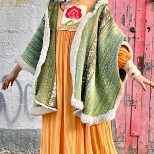 Puede incluir: Conjunto femenino con un vestido largo y fluido de color naranja, combinado con un poncho tejido verde y blanco con ribete de piel sintética blanca. El vestido tiene un escote cuadrado con bordado floral. Botas de cuero marrón completan el look.