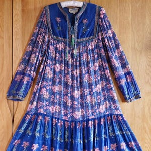 Puede incluir: Un vestido de manga larga con estampado floral azul y rosa, falda fruncida y cuello bordado. El vestido tiene un lazo en el cuello y una borla.