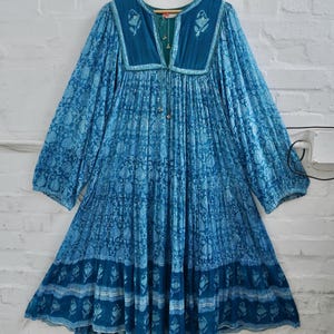 May include: A long-sleeved blue dress featuring a deep V-neckline with a tie closure, adorned with a repeating floral block print pattern in varying shades of blue and white, with a gathered bodice and a tiered, flared skirt silhouette.