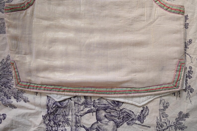 May include: A white fabric with a green and red trim. The fabric has a subtle sheen and is slightly wrinkled. The fabric is lying on a white surface with a floral pattern.