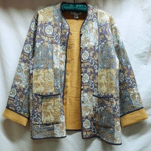 May include: A quilted jacket with a floral pattern in shades of purple, gold, and gray. The jacket has a mandarin collar, two front pockets, and a mustard yellow lining. The cuffs and trim are also mustard yellow.