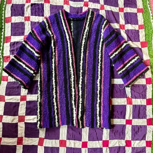 May include: A purple, black, and white striped cardigan with a fuzzy texture. The cardigan has a V-neck and long sleeves.