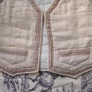 May include: A white quilted vest with green, red, and pink trim. The vest has two pockets on the front and a V-neck.